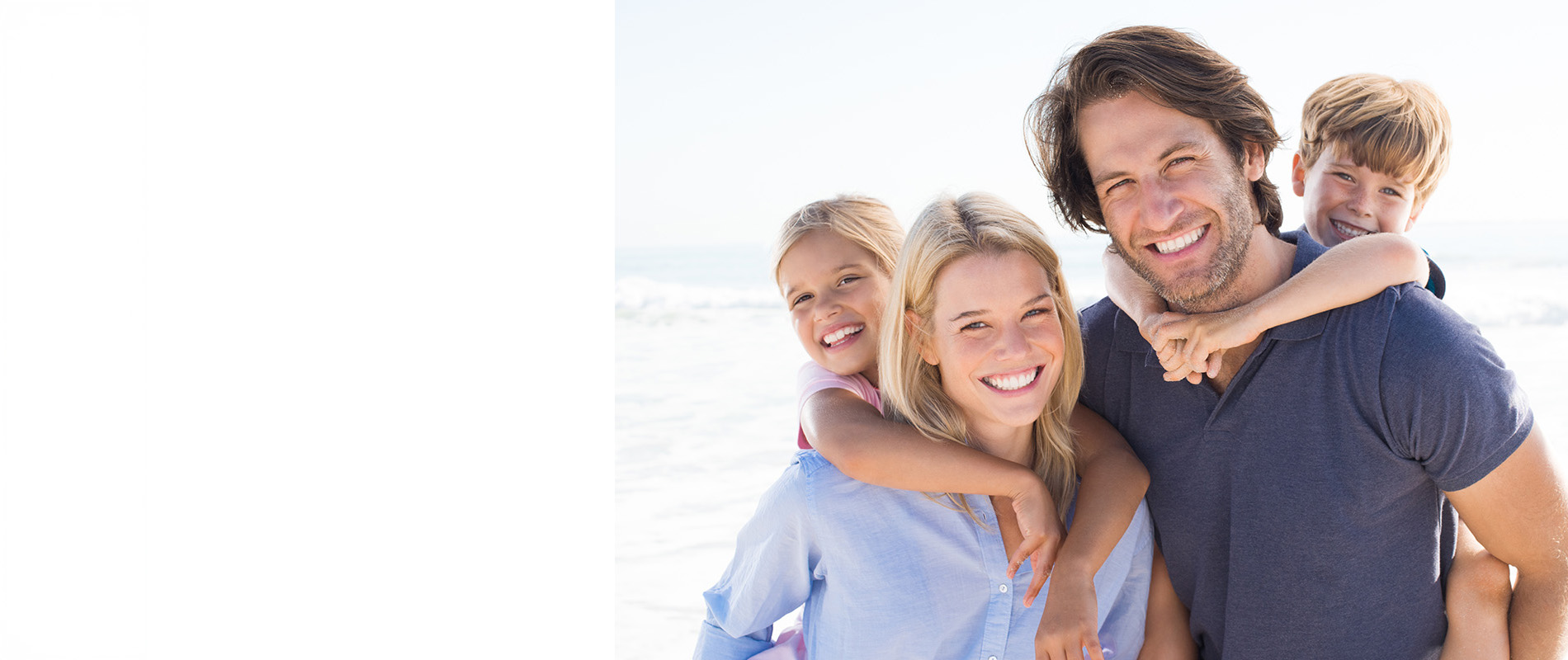 The image depicts a family of four on a beach, with the father standing in front of his wife and two children, all smiling and looking towards the camera.