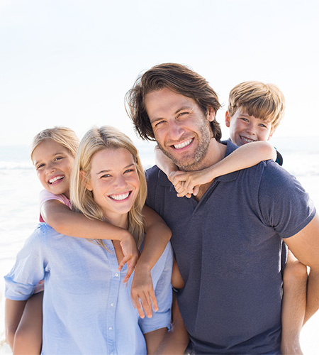 The image shows a family of four standing on a beach with clear skies, the father holding two children while smiling at the camera, and the mother behind them also smiling.