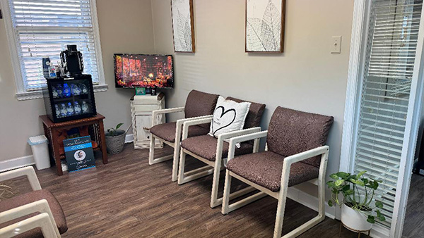 The image shows an interior room with furniture and decor, including chairs, a couch, a television, and a coffee table. There are framed pictures on the wall and a window with blinds partially drawn.