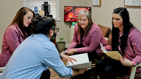 Four women sitting around a table with a man standing over them, all engaged in conversation, possibly discussing business matters.