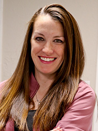 The image shows a woman standing against a blurred background with her arms crossed, wearing a pink jacket over a black top, smiling at the camera.