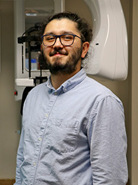 The image shows a man standing indoors, wearing glasses, a blue shirt, and a light-colored pants. He has facial hair and appears to be smiling at the camera. Behind him, there is medical equipment, including a large white machine with a screen displaying text, which suggests that he may be in a healthcare setting.