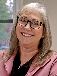 The image depicts a woman with glasses, smiling at the camera, wearing a pink jacket and black eyeglasses, against a blurred background of an indoor setting.