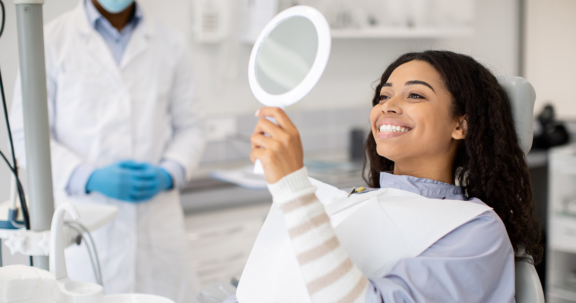 A woman in a dental chair with a dentist behind her, smiling at the camera while holding a mirror.