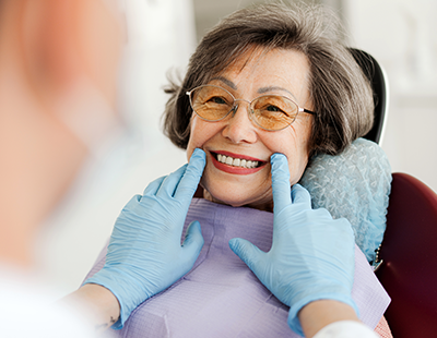 This is a photograph featuring an elderly woman seated in a dental chair with her eyes closed, smiling broadly while undergoing dental treatment. A dental hygienist stands behind her, holding a mirror and adjusting the woman s mouth with one hand, while the other hand gently holds her chin. The woman wears glasses, has gray hair, and is wearing a blue surgical mask. She is also wearing a purple neck brace. There are no visible texts on the image.