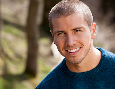 A man with short hair and a beard, smiling at the camera, wearing a blue sweater, standing outdoors in front of trees.
