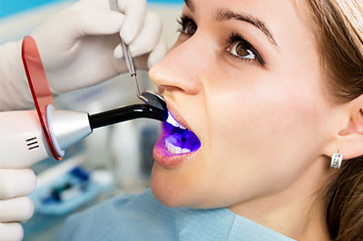 The image shows a woman sitting in a dental chair with her mouth open wide while receiving dental care, with a dentist using a device to examine her teeth.