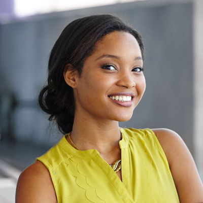 The image features a smiling woman with dark hair, wearing a yellow top and a necklace, posing against a blurred background with a neutral expression.